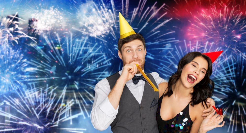 birthday, celebration and holidays concept - happy couple with party blowers and caps having fun over firework background. happy couple with party blowers having fun