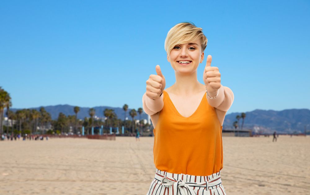 travel, tourism and summer holidays concept - happy smiling young woman showing thumbs up over venice beach background in california. happy smiling young woman showing thumbs up