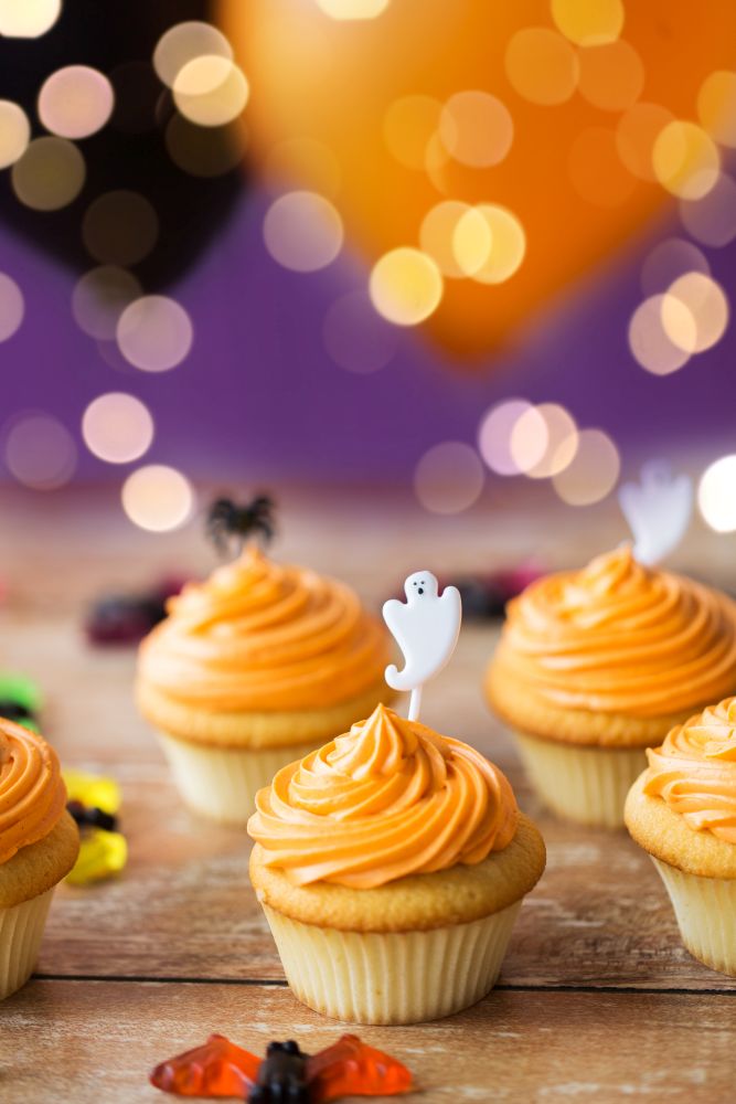 food, baking and holidays concept - cupcakes or muffins with halloween party ghost decoration on wooden table over lights. cupcakes with halloween decoration on table