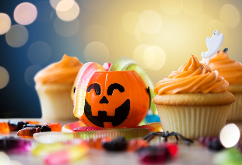 halloween and holidays concept - cupcakes or frosted muffins with party decorations and candies on wooden table over lights. halloween party decorated cupcakes on wooden table