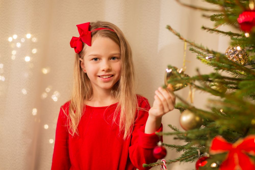 christmas, holidays and childhood concept - happy girl in red clothes decorating natural fir tree. happy girl in red decorating christmas tree