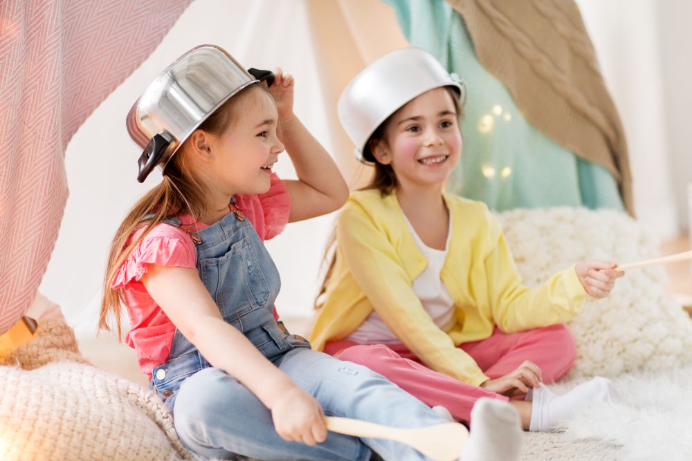 childhood and hygge concept - happy little girls with cooking pots playing in kids tent at home. girls with kitchenware playing in tent at home