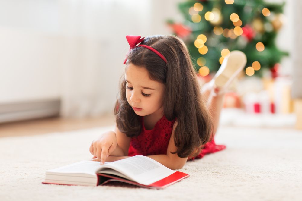 christmas, holidays and childhood concept - happy little girl lying on floor and reading book at home. happy girl reading book at home on christmas