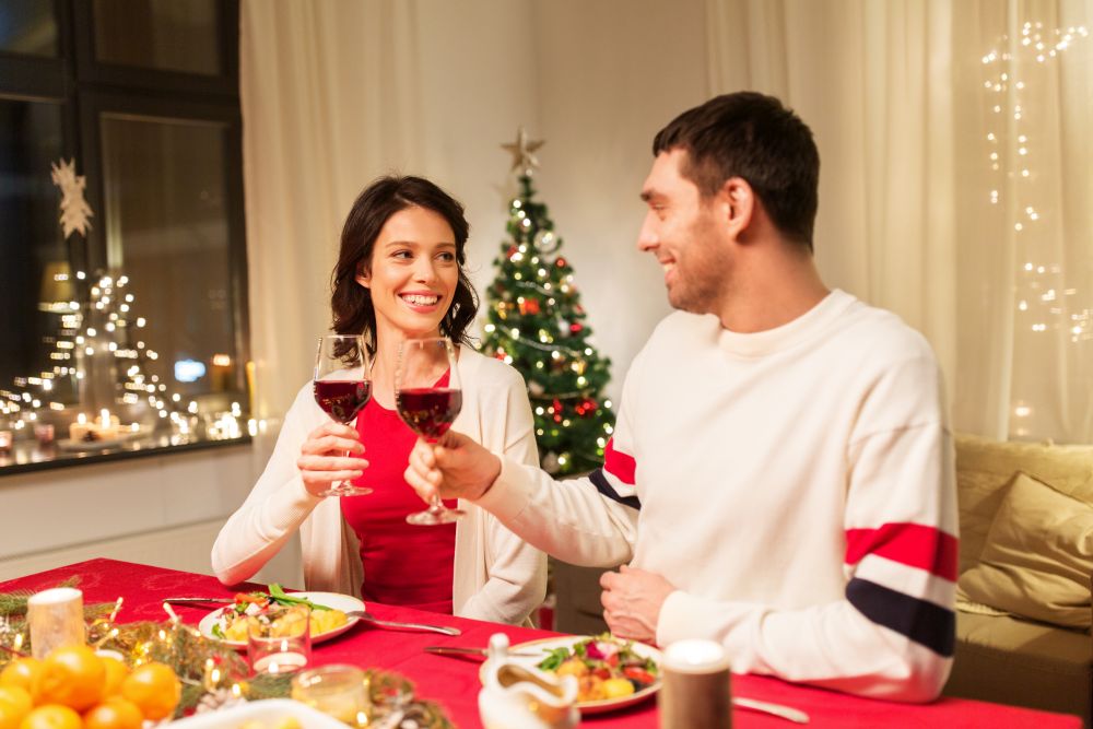 holidays, family and celebration concept - happy couple having christmas dinner at home and drinking red wine and clinking glasses. happy couple drinking red wine at christmas dinner