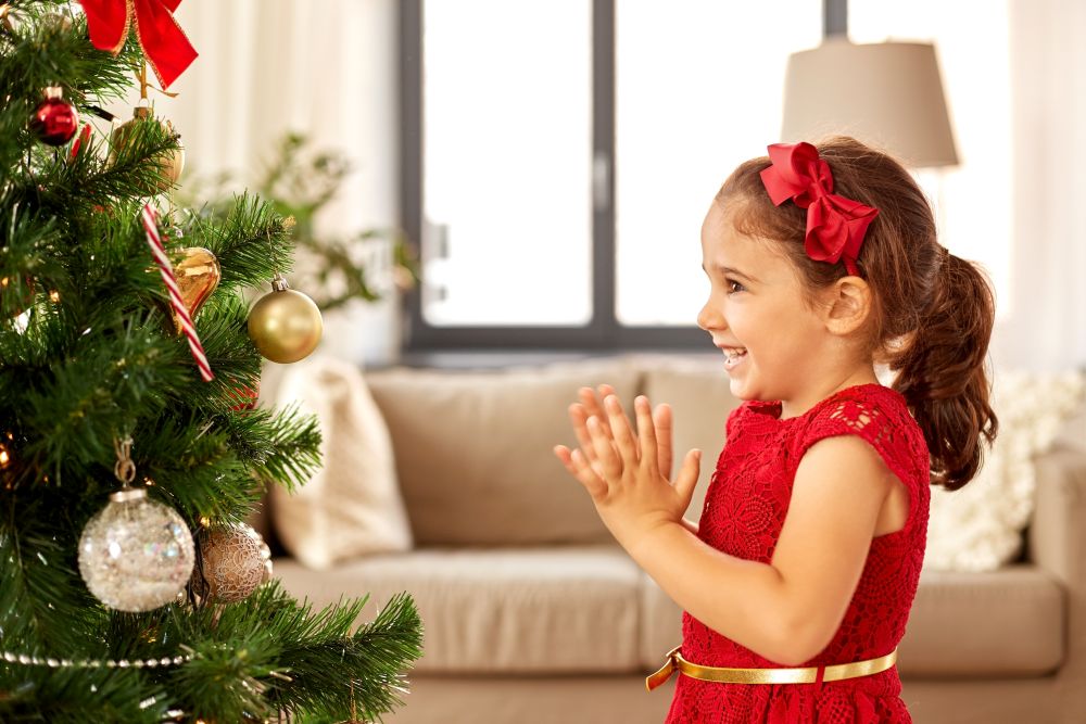 winter holidays and people concept - happy little girl decorating christmas tree at home. little girl decorating christmas tree at home