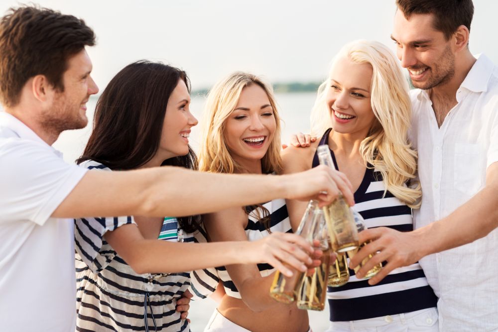 friendship, summer holidays and people concept - group of happy friends in striped clothes drinking non alcoholic beer on beach. happy friends drinking non alcoholic beer on beach