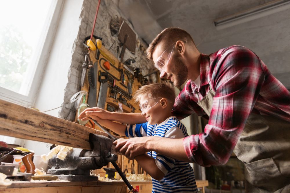 family, carpentry, woodwork and people concept - happy father and little son with rasp grinding wood plank at workshop. father and son with rasp working at workshop