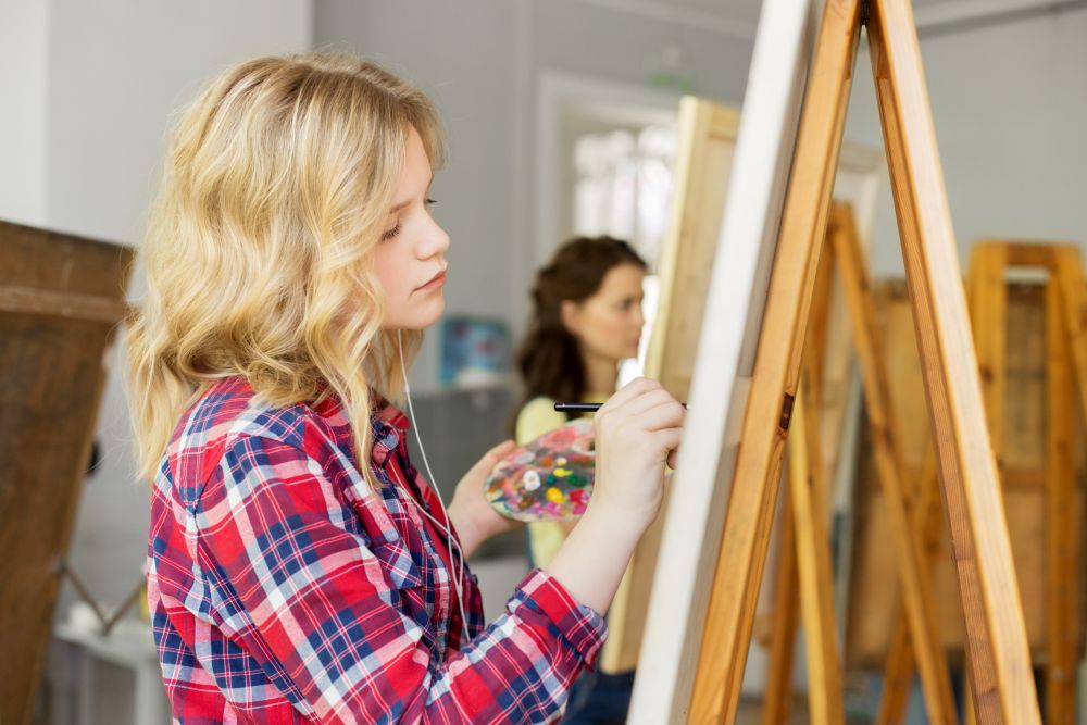 art school, creativity and people concept - teenage girl in earphones with easel, palette and brush painting and listening to music at studio. girl with easel painting at art school studio