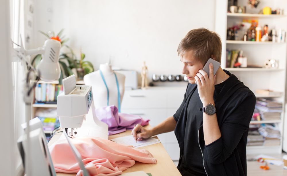 people, clothing and tailoring concept - fashion designer calling on smartphone and making notes at studio. fashion designer calling on smartphone at studio