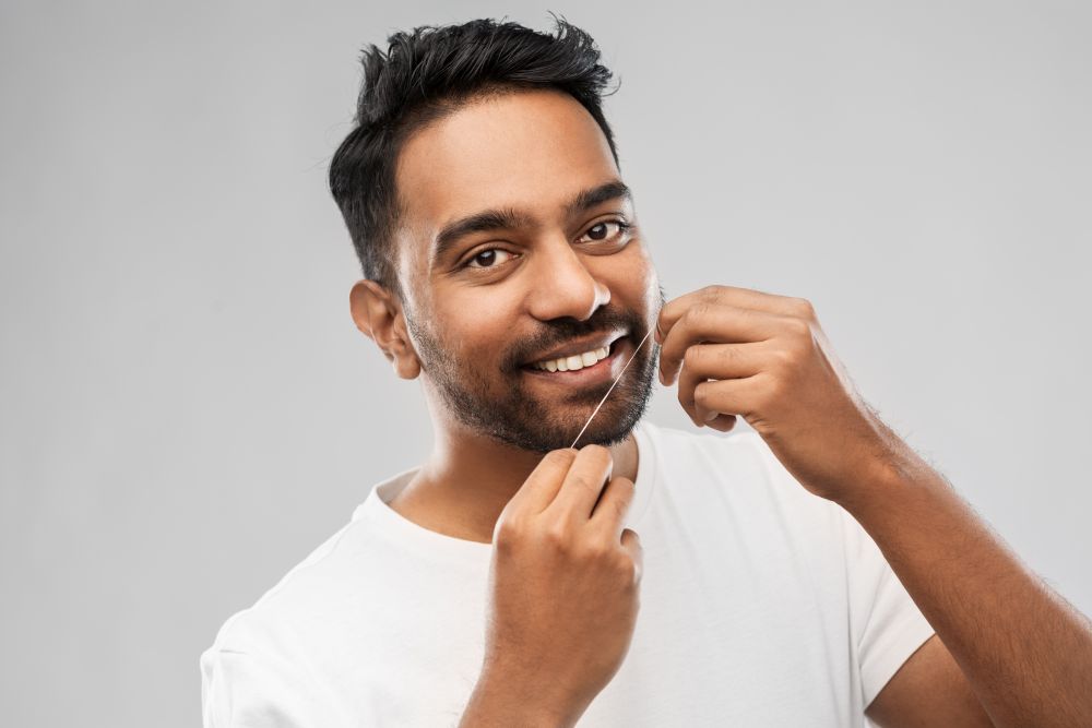 oral care, hygiene and people concept - smiling young indian man with dental floss cleaning teeth over gray background. indian man with dental floss cleaning teeth