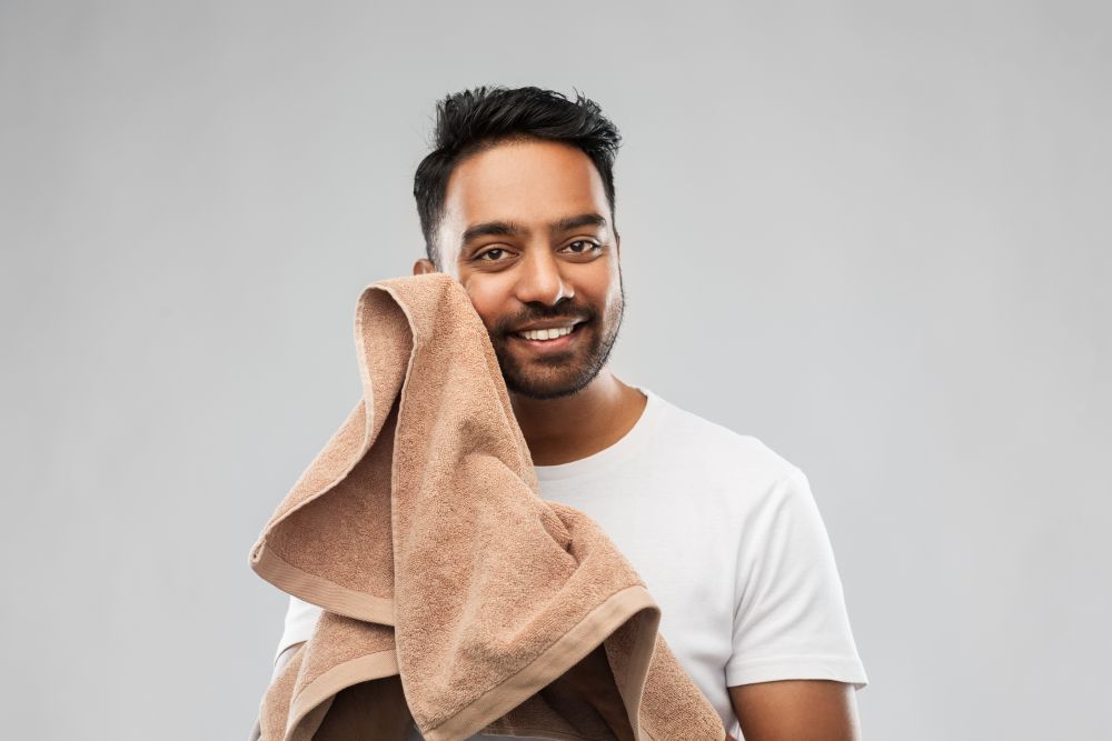 grooming and people concept - smiling indian man with bath towel drying his face over grey background. smiling indian man with towel over grey background