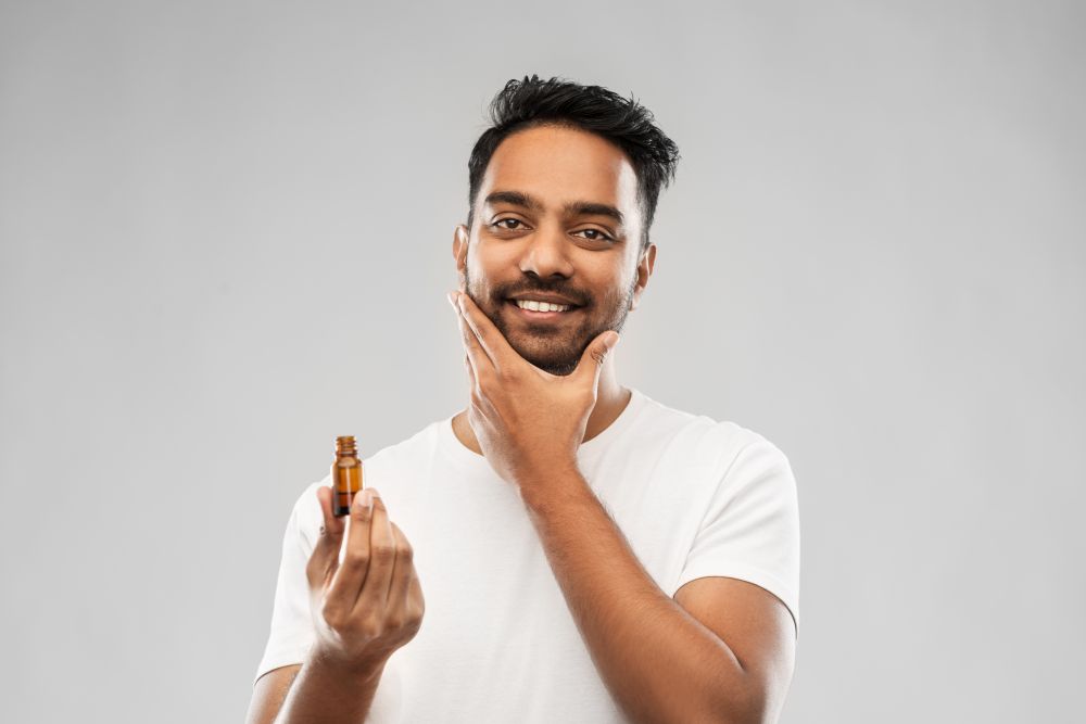 grooming and people concept - smiling young indian man applying lotion or beard oil over gray background. smiling indian man applying grooming oil to beard