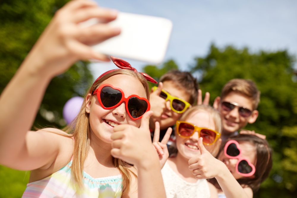 childhood, friendship and technology concept - happy kids in sunglasses taking selfie and showing thumbs up at summer park. kids taking selfie and showing thumbs up at park