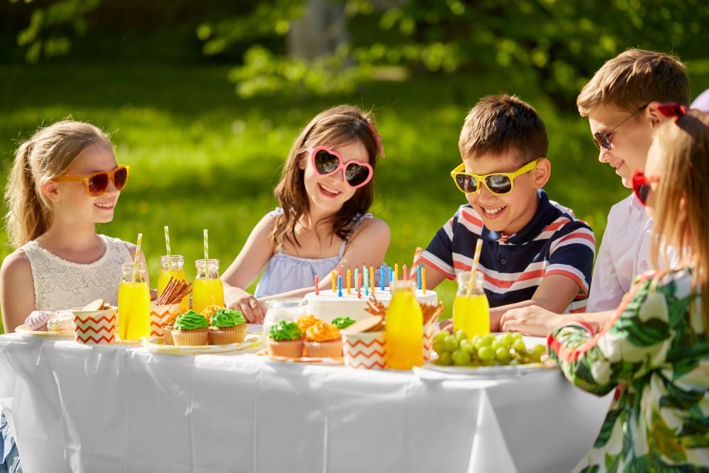 holidays, childhood and celebration concept - happy kids with candles on birthday cake sitting at table at summer garden party. happy kids with cake on birthday party in summer