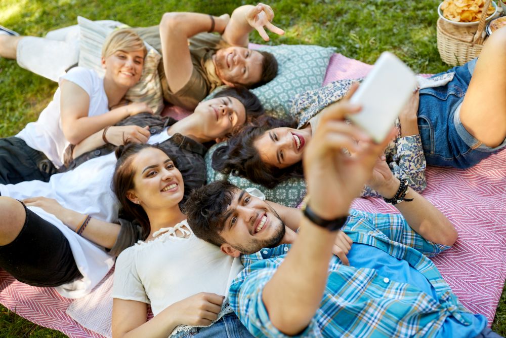 friendship, leisure, technology and people concept - group of friends chilling on picnic blanket at summer park and taking selfie by smartphones. friends taking selfie on picnic at summer park