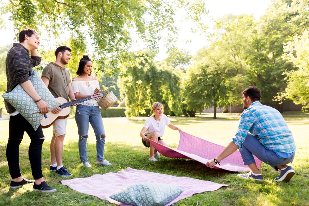 friendship and leisure concept - group of happy friends with guitar, basket and blanket arranging place for picnic at summer park. friends arranging place for picnic at summer park