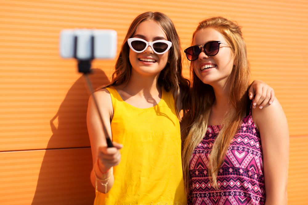 fashion, leisure and people concept - smiling teenage girls taking picture by smartphone on selfie stick outdoors in summer. teenage girls taking picture by selfie stick