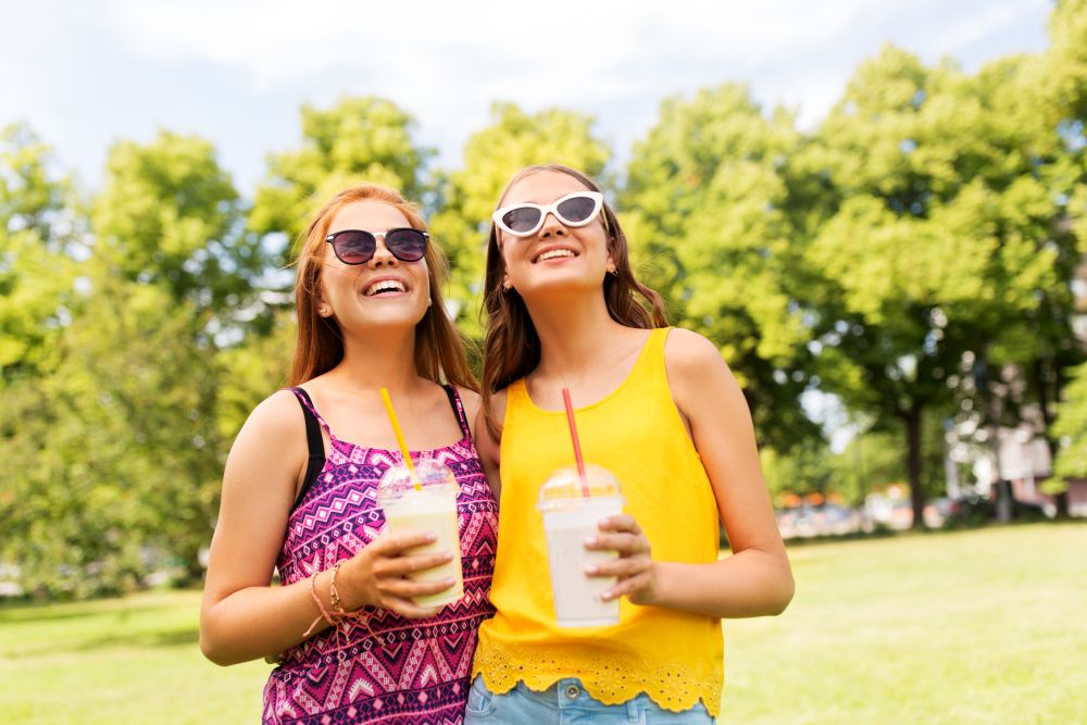 leisure and friendship concept - happy smiling teenage girls or friends with milk shakes or smoothie drinks at summer summer park. teenage girls with milk shakes at summer park