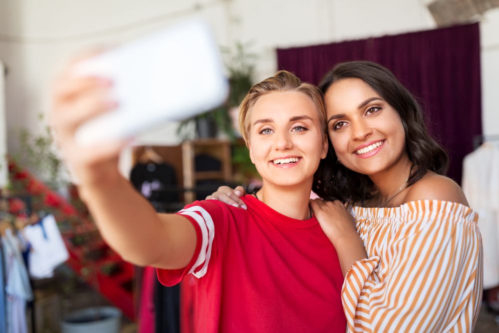 sale, shopping and technology concept - happy female friends taking selfie by smartphone at vintage clothing store. female friends taking selfie at clothing store