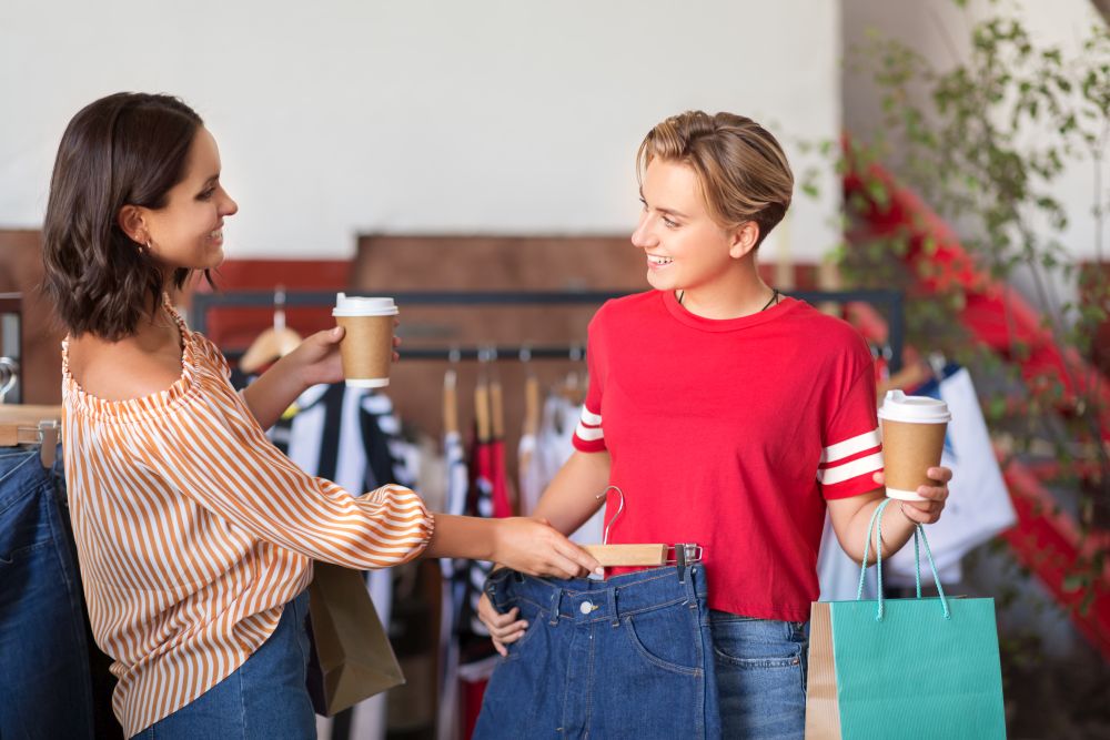 sale, shopping, fashion and people concept - happy young women with takeaway coffee cups choosing clothes at vintage clothing store. happy women with coffee at vintage clothing store