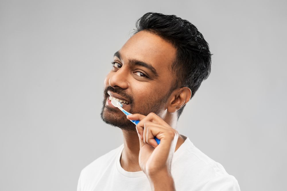 oral care, dental hygiene and people concept - smiling young indian man with toothbrush cleaning teeth over gray background. indian man with toothbrush cleaning teeth