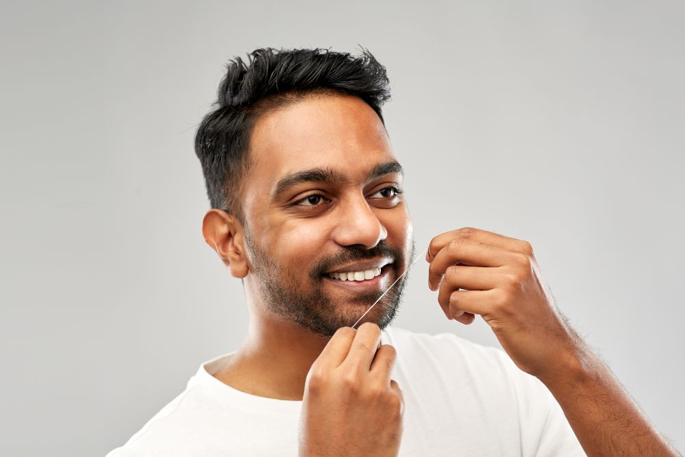 oral care, hygiene and people concept - smiling young indian man with dental floss cleaning teeth over grey background. indian man with dental floss cleaning teeth