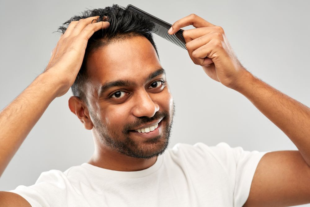 grooming, hairstyling and people concept - smiling young indian man brushing hair with comb over grey background. happy indian man brushing hair with comb