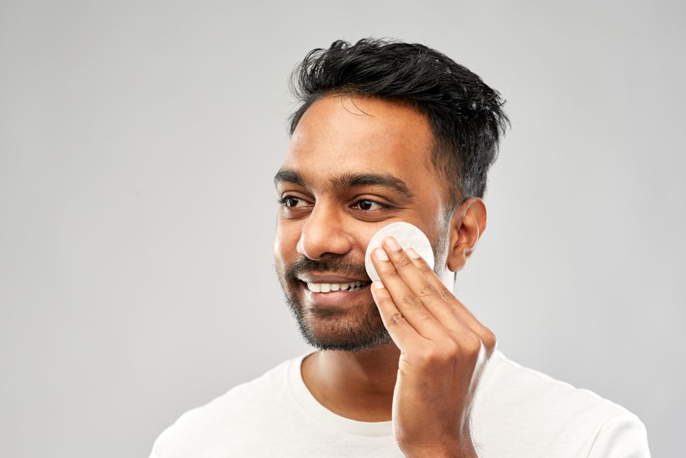 grooming, skin care and people concept - smiling young indian man cleaning his face with cotton pad over grey background. smiling indian man cleaning face with cotton pad