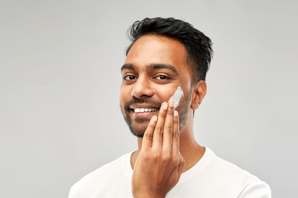 grooming, skin care and people concept - smiling young indian man applying cream to face over grey background. happy indian man applying cream to face