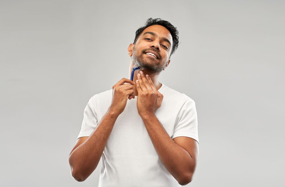 grooming and people concept - young indian man shaving beard with manual razor blade over grey background. indian man shaving beard with razor blade