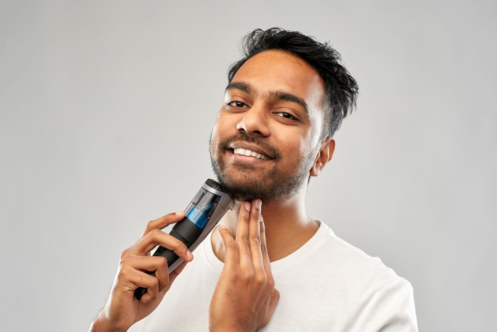 grooming, technology and people concept - smiling indian man shaving beard with trimmer over grey background. smiling indian man shaving beard with trimmer