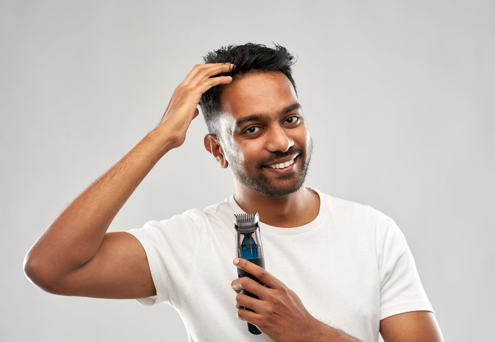 grooming and people concept - smiling young indian man with trimmer touching his hair over grey background. smiling indian man with trimmer touching his hair