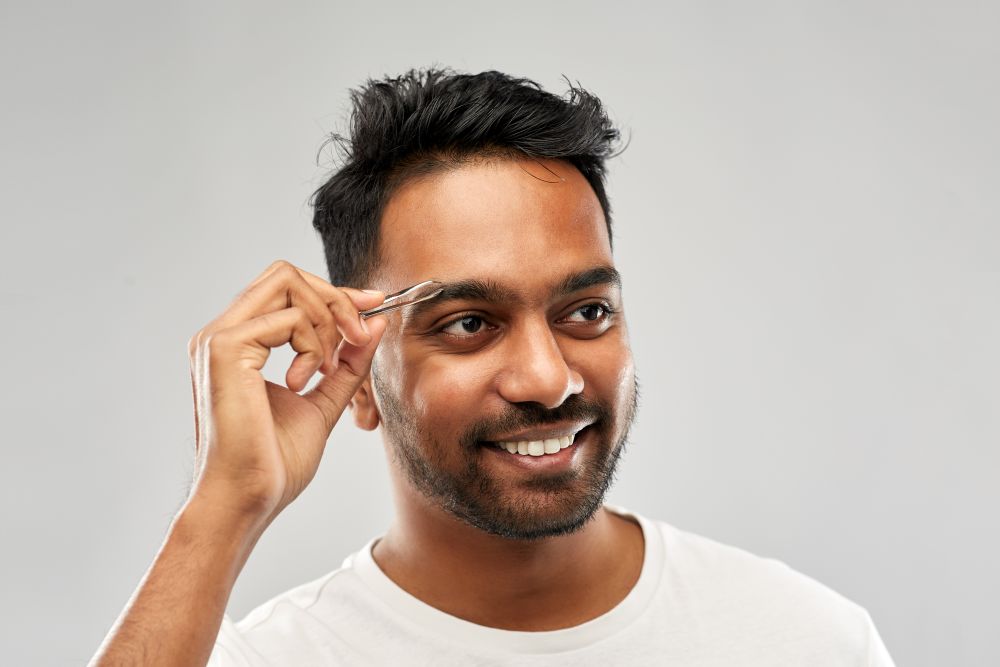 grooming and people concept - smiling young indian man with tweezers tweezing eyebrow hair over grey background. indian man with tweezers tweezing eyebrow hair