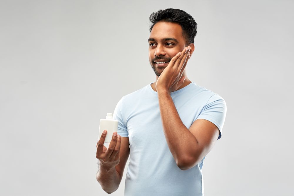 grooming, skin care and people concept - smiling young indian man applying lotion to face over grey background. happy indian man applying lotion to face
