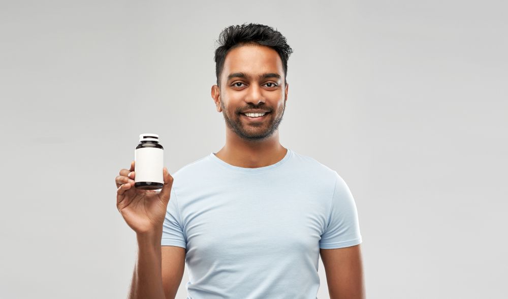 medicine, healthcare and people concept - smiling young indian man holding jar with some drug over grey background. indian man with medicine over grey background