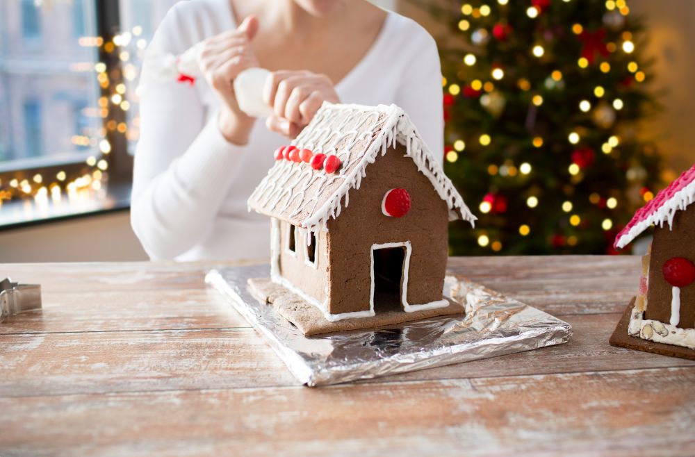 cooking, holidays and people concept - woman with pastry bag making gingerbread houses at home over christmas tree lights background. woman making gingerbread houses on christmas