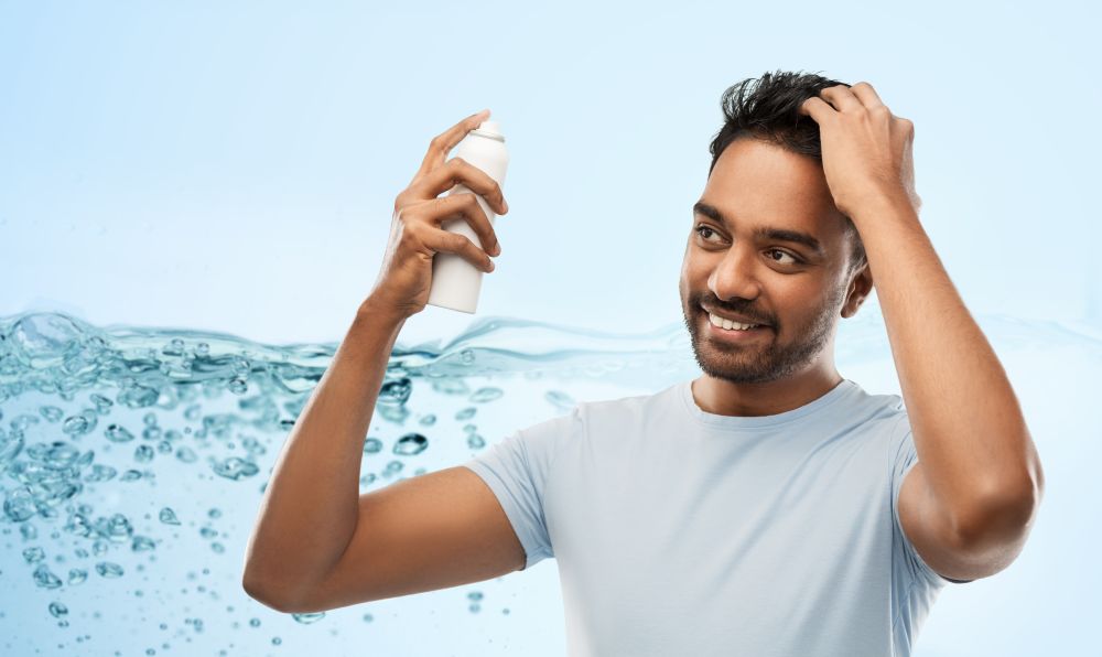 grooming, hairstyling and people concept - happy smiling indian man applying hair spray over blue background with air bubbles or water splash. smiling indian man applying hair spray over gray
