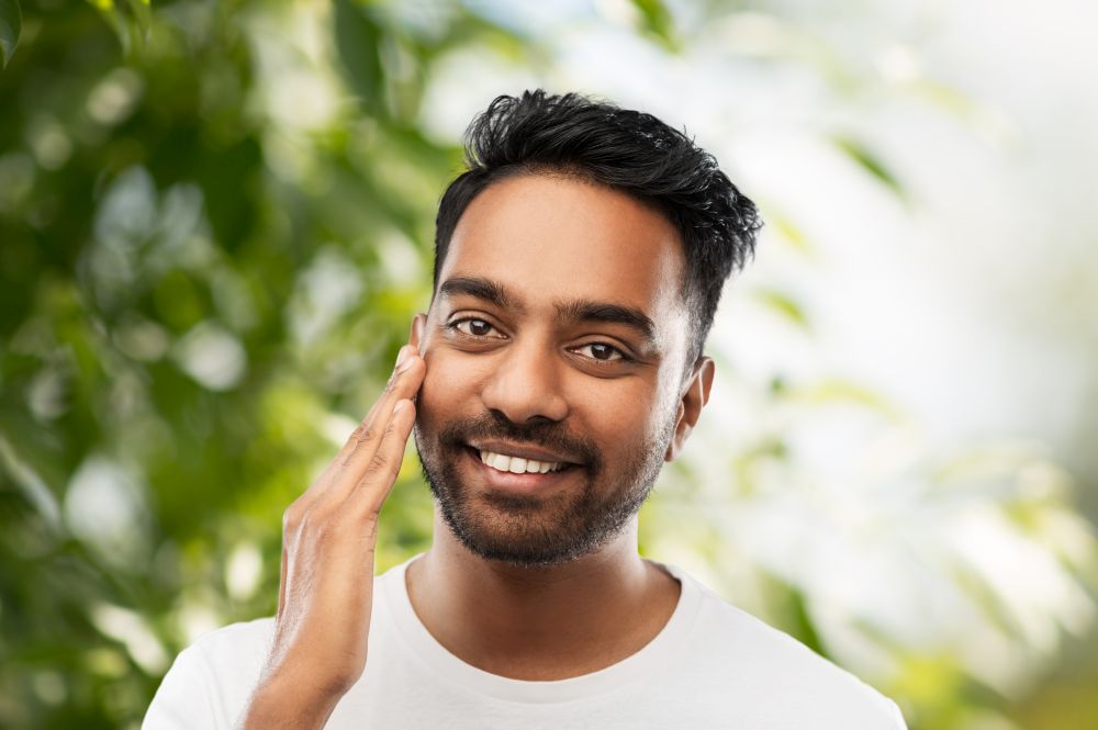 grooming, skin care and people concept - smiling young indian man touching his face over green natural background. smiling indian man touching his face