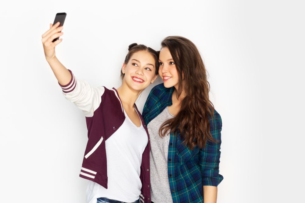 technology and friendship concept - happy smiling pretty teenage girls taking selfie by smartphone over white background. happy teenage girls taking selfie by smartphone