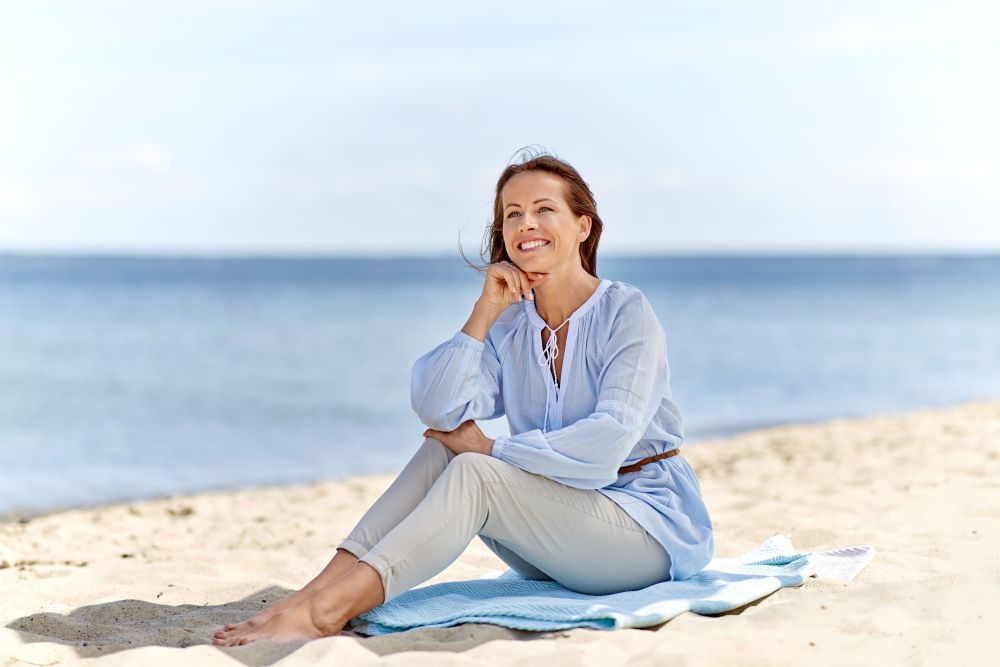 summer, people and leisure concept - happy smiling woman sitting on beach towel. happy woman sitting on summer beach