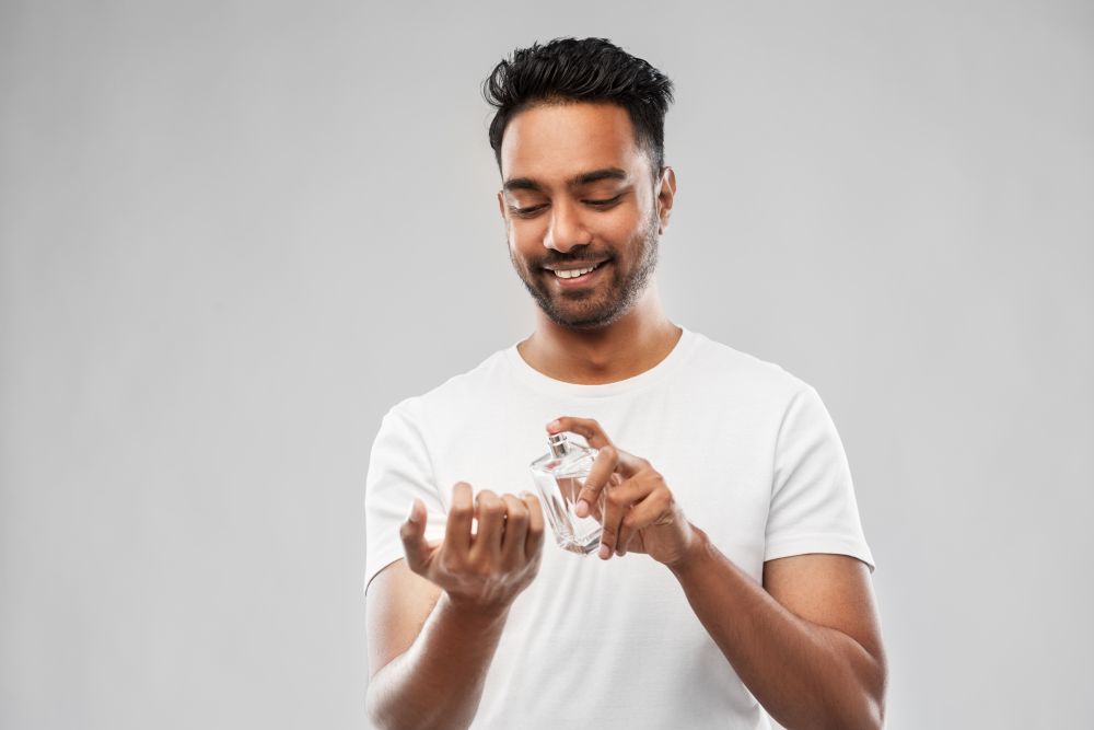 male perfumery, grooming and people concept - happy smiling young indian man spraying perfume to his wrist over gray background. happy indian man with perfume over gray background