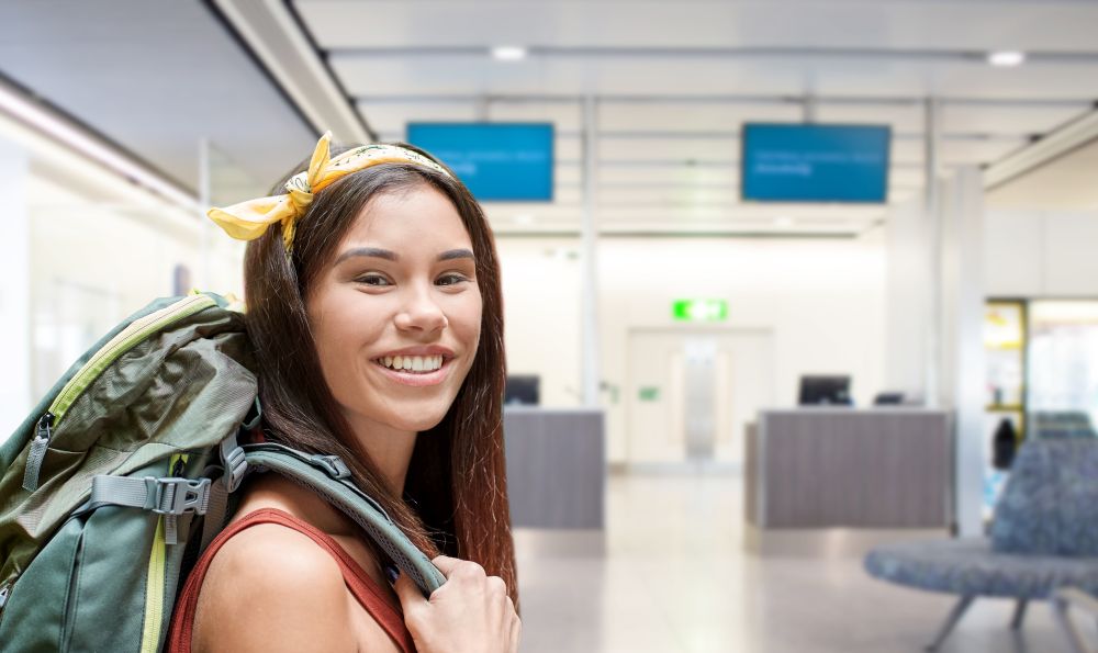 travel, tourism and people concept - smiling young woman with backpack over airport terminal background. young woman with backpack over airport terminal