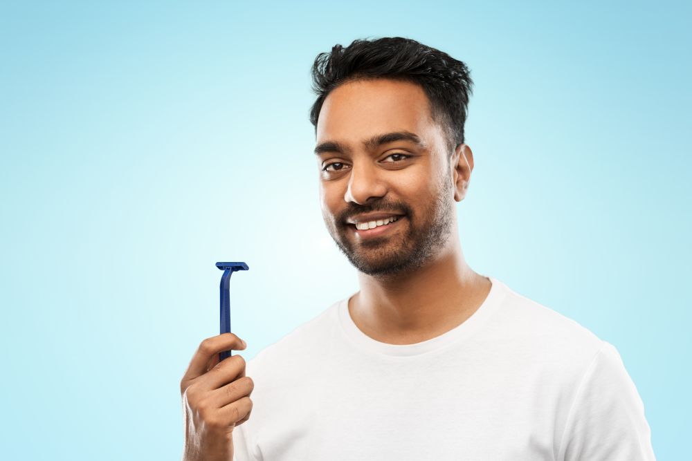 beard shaving, grooming and people concept - smiling indian man with manual razor blade over blue background. indian man shaving beard with razor blade