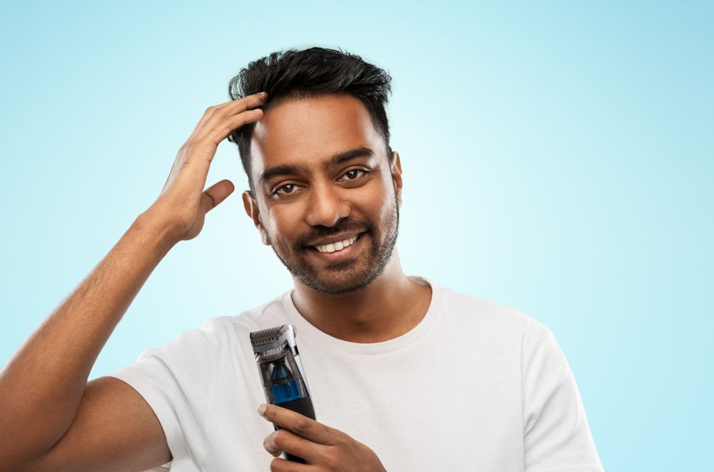 grooming and people concept - smiling young indian man with trimmer touching his hair over blue background. smiling indian man with trimmer touching his hair