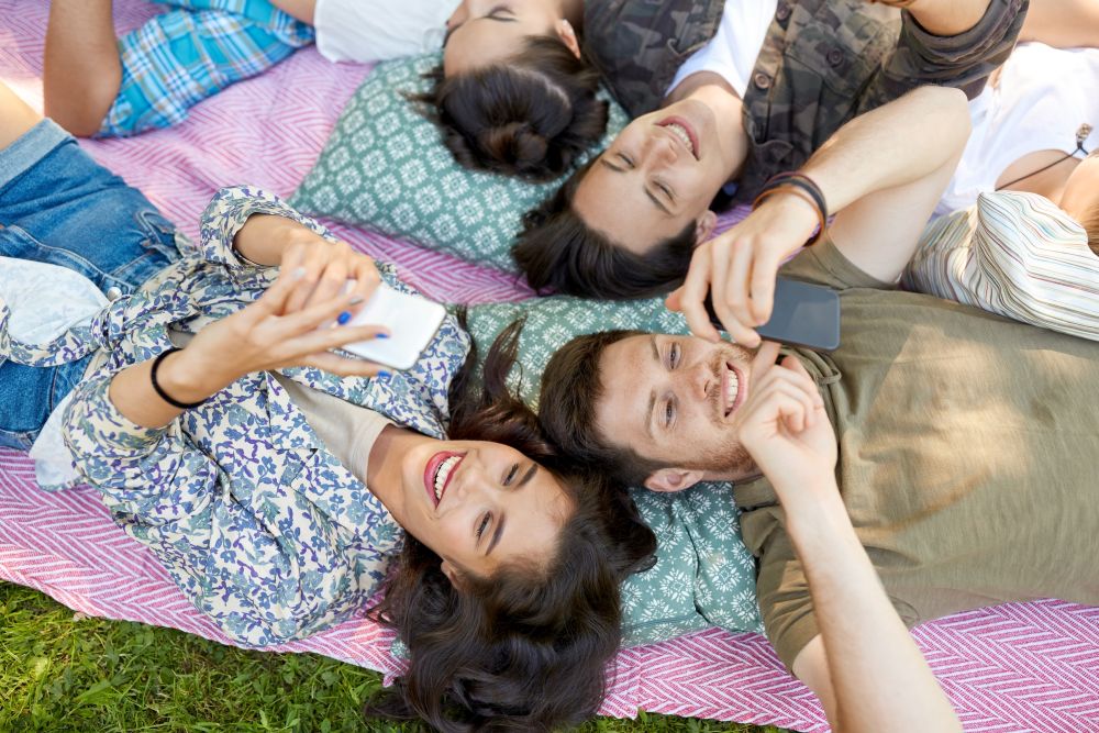 friendship, leisure, technology and people concept - group of friends with smartphones chilling on picnic blanket at summer park. friends with smartphones chilling at summer park
