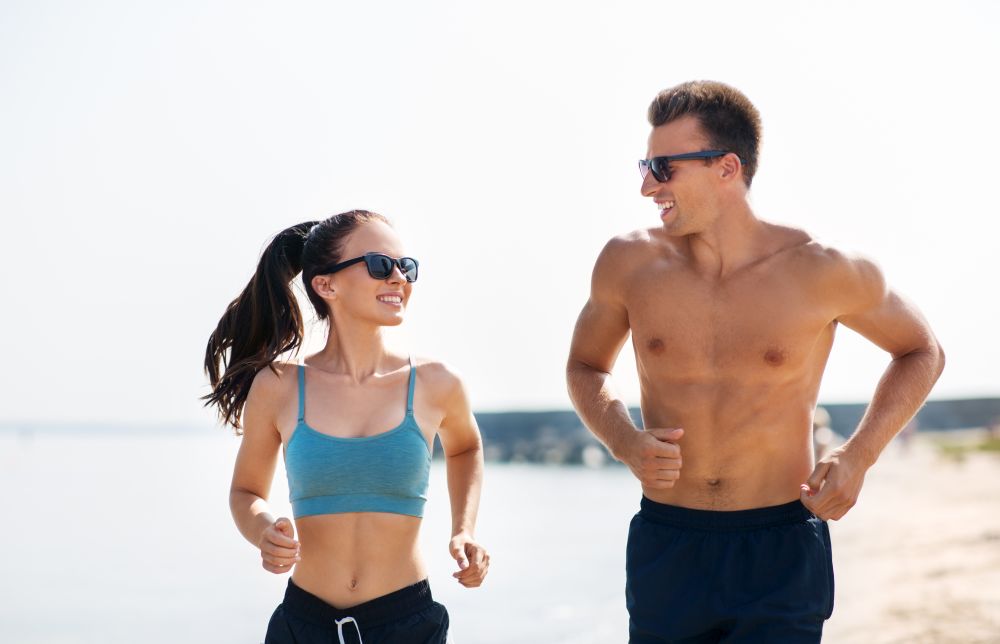 fitness, sport and lifestyle concept - happy couple in sports clothes and sunglasses running along summer beach. couple in sports clothes running along on beach