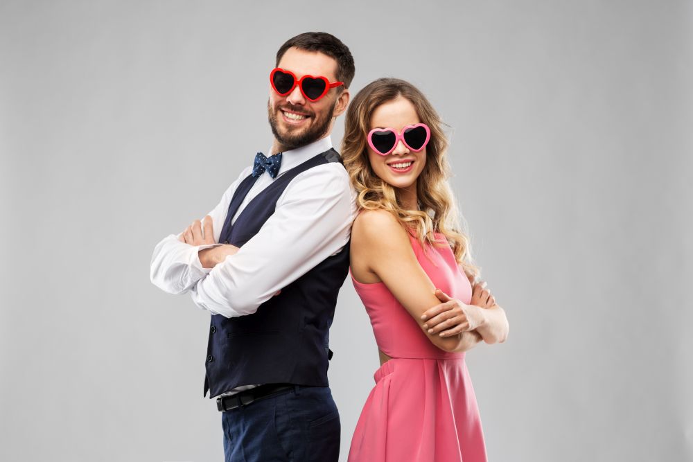 valentines day, love and people concept - happy couple in heart-shaped sunglasses over grey background. happy couple in heart-shaped sunglasses