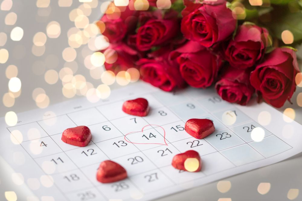 valentines day and holidays concept - close up of calendar sheet with marked 14th february date, heart shaped chocolate candies and red roses over festive lights. close up of calendar, heart, candies and red roses