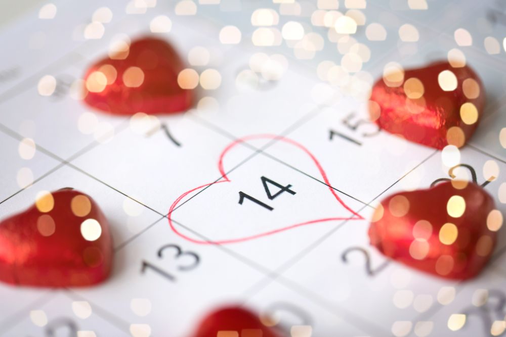 valentines day and holidays concept - close up of calendar sheet with marked 14th february date and red heart shaped chocolate candies over festive lights. close up of calendar and heart shaped candies