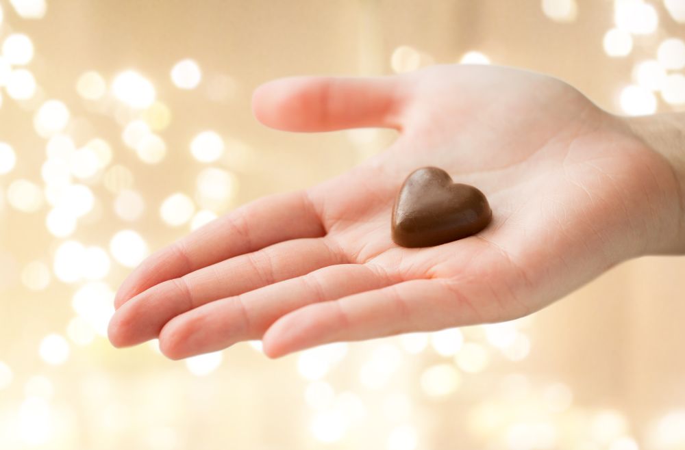 valentines day, sweets and confectionery concept - close up of hand holding heart shaped chocolate candy over festive lights background. close up of hand with heart shaped chocolate candy